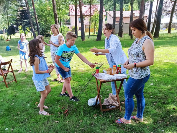 Children and their guardians stand on the grass at a snack table. One of the boys puts a sausage on a metal skewer