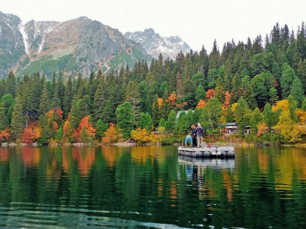 A mountain pond surrounded by a forest in autumn colours and mountains. A platform with three people floats on its surface