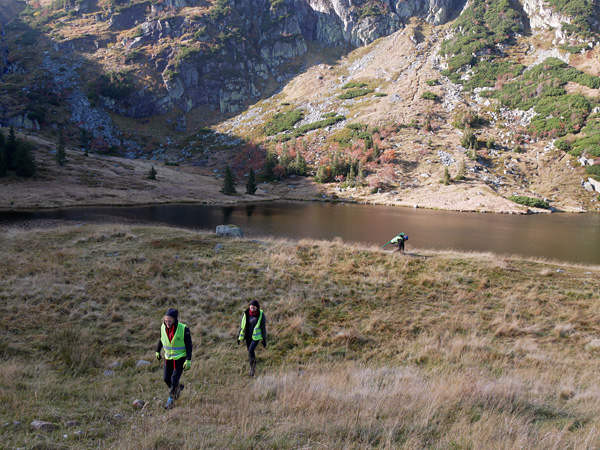 Three people in reflective vests walk across a meadow with a mountain pond in the background