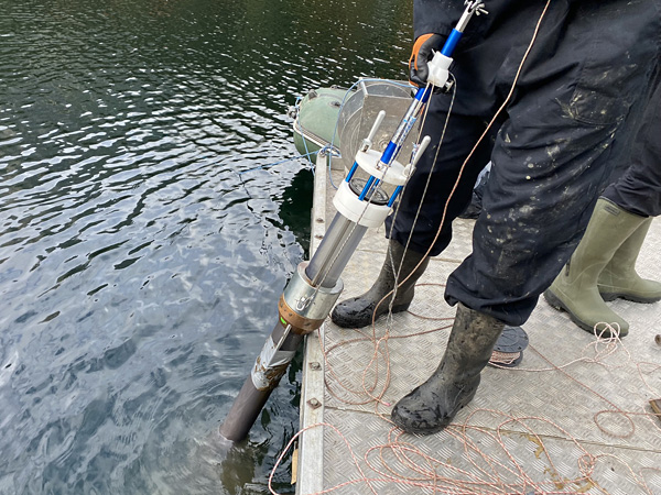 A person in a muddy wetsuit stands on the edge of a platform and holds a long device submerged in the water