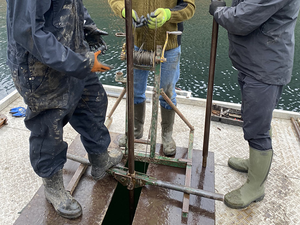 Three people seen from the waist down in work clothes stand at a winch mounted in the middle of a floating platform