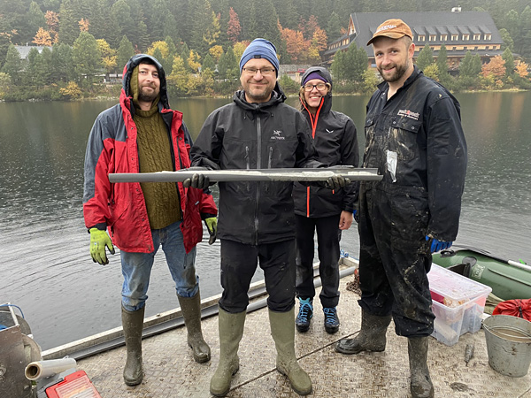 Three men and a woman stand on a floating platform. One of the men proudly displays a long sample of sediment