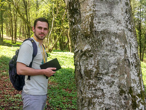 A young man with a notebook in his hand stands in a sunlit forest. Next to him is a tree trunk covered with spots of lichen