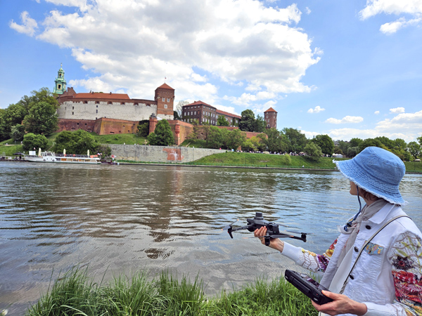 Woman in a hat holding a drone stands by a wide river; a castle is visible on the opposite bank