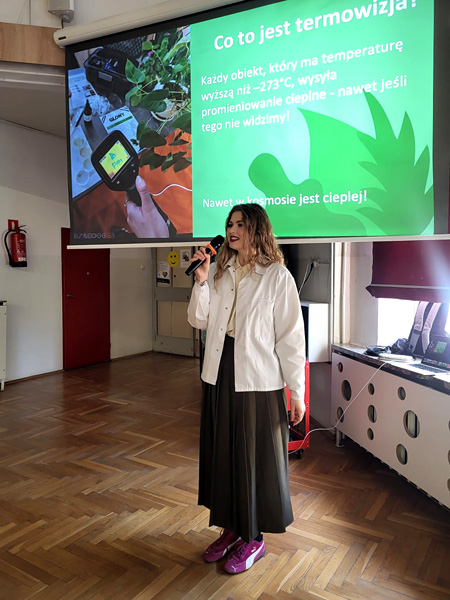 A young woman stands in front of a presentation screen, smiling as she speaks into a microphone