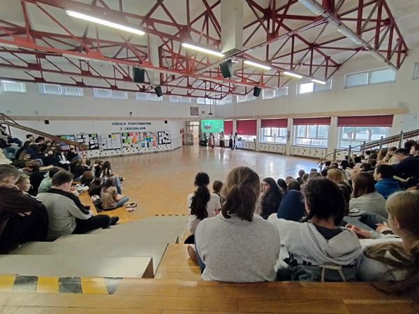 Large gym hall; a crowd of students sits on steps, watching a presentation in the distance