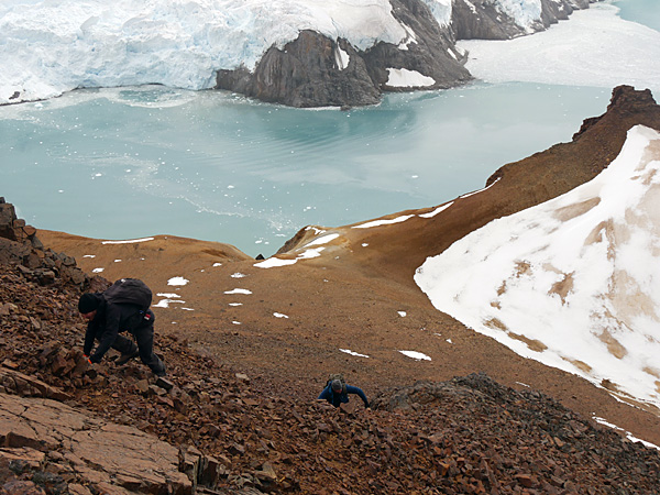 A view down a steep, brown, rocky slope at two people climbing. In the background, a glacier and a frozen surface of water